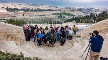 A photographer capturing a photo of a group of tourists on the Mount Olives with the Old City of Jerusalem in the background. Image via Shutterstock.com