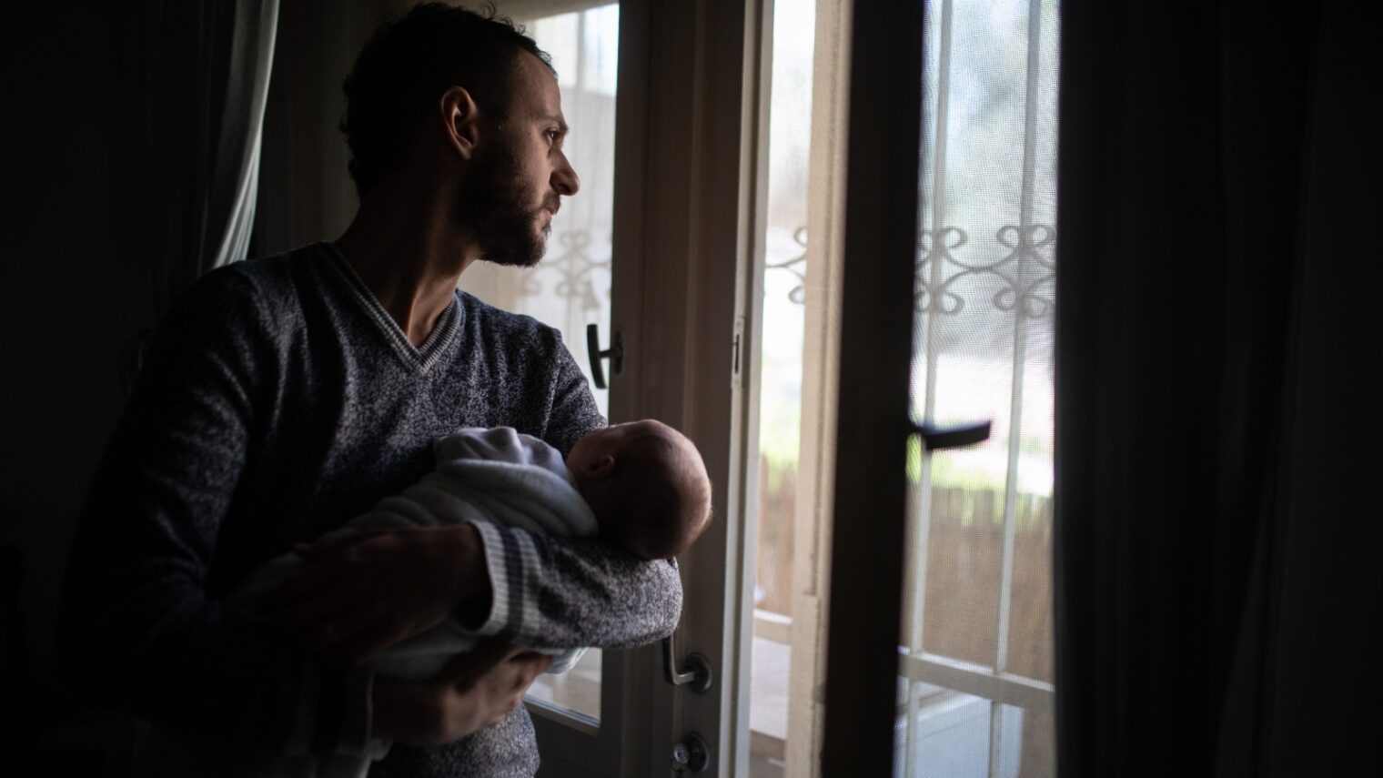 A Jerusalem father holds his baby as he looks out the window during a nationwide partial lockdown, April 3, 2020. Photo by Hadas Parush/Flash90
