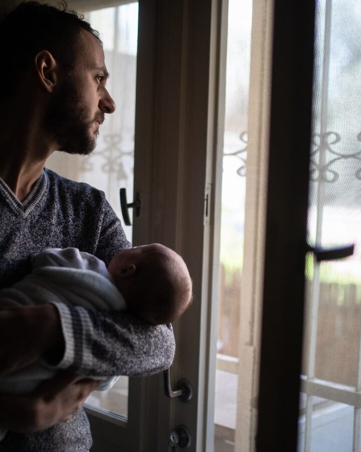 A Jerusalem father holds his baby as he looks out the window during a nationwide partial lockdown, April 3, 2020. Photo by Hadas Parush/Flash90